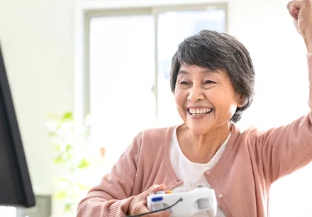 コントローラーを手にモニターの前でガッツポーズをする高齢女性（利用イメージ）