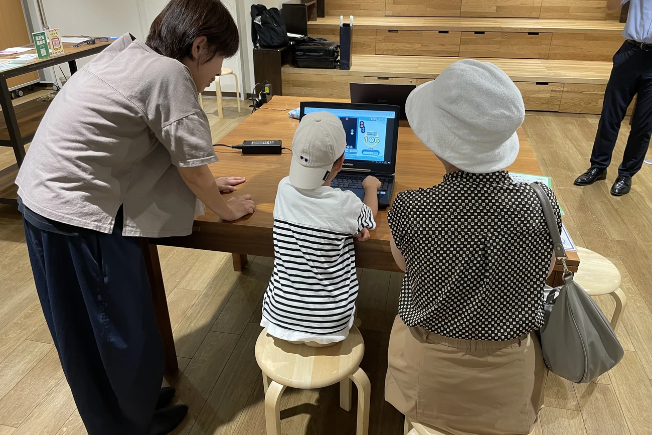 並んで座った参加者2名と、机のそばに立って見守る運営スタッフ